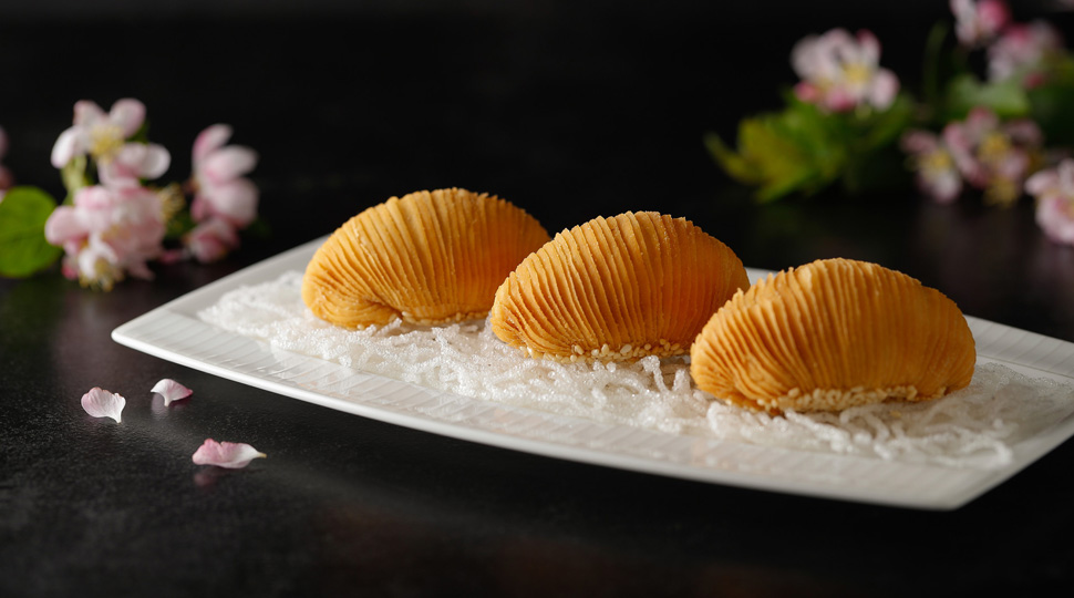 Three fried Australian wagyu beef puffs on a white rectangular plate set on a black background with springs of pink flower blossoms