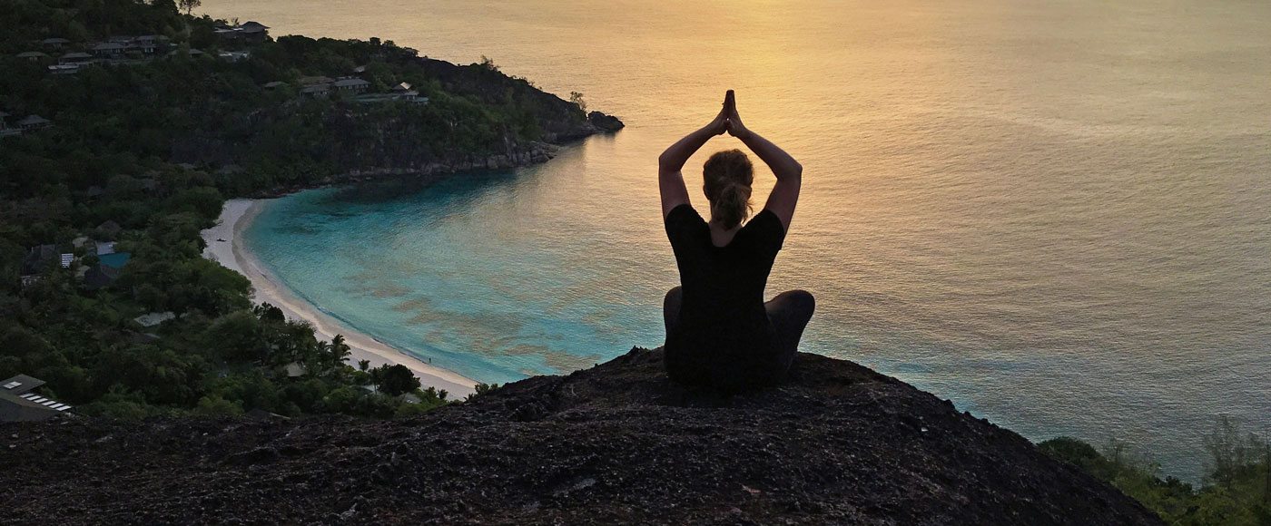 A woman sits a top of peak at sunset overlooking the ocean