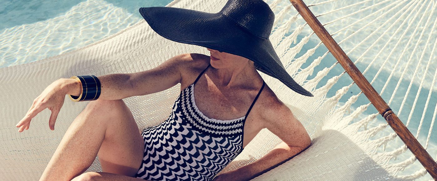 Woman in a black and white abstract print swimsuit and large black sunhat lounges in a hammock