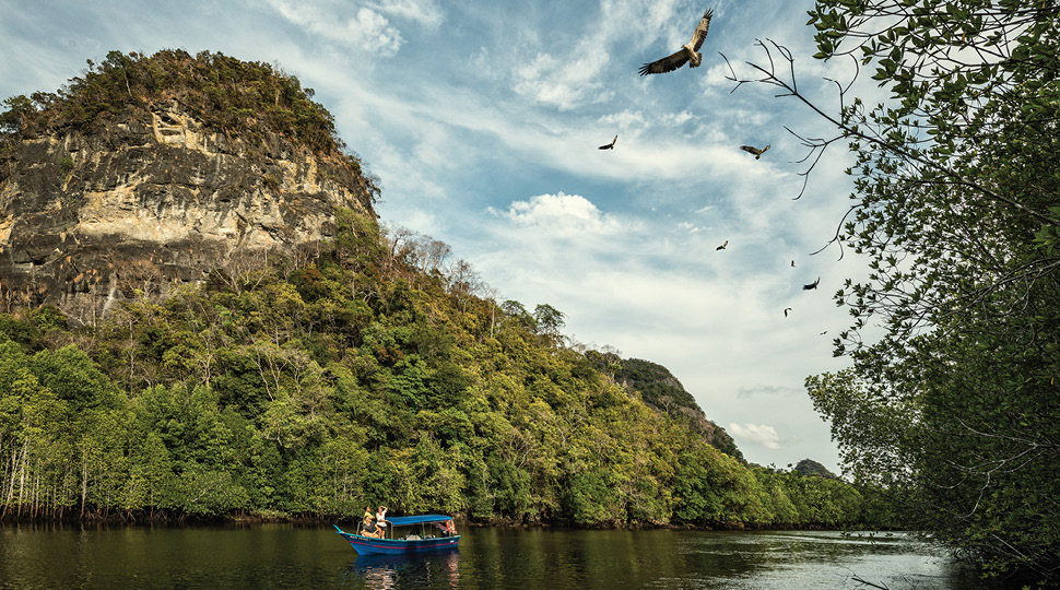 People on small blue boat on river under small rocky mountain, with eagles flying above