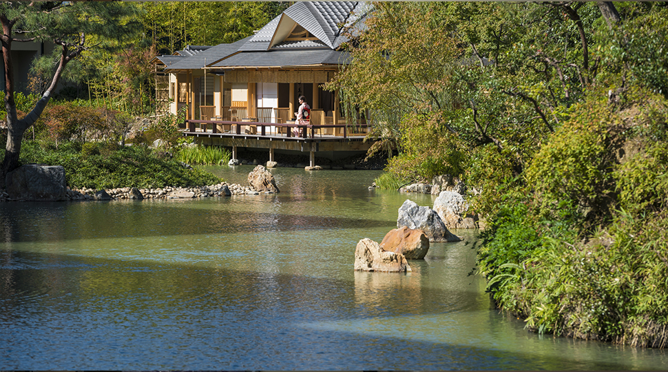 View across pond to bungalow where woman in robe sits on patio