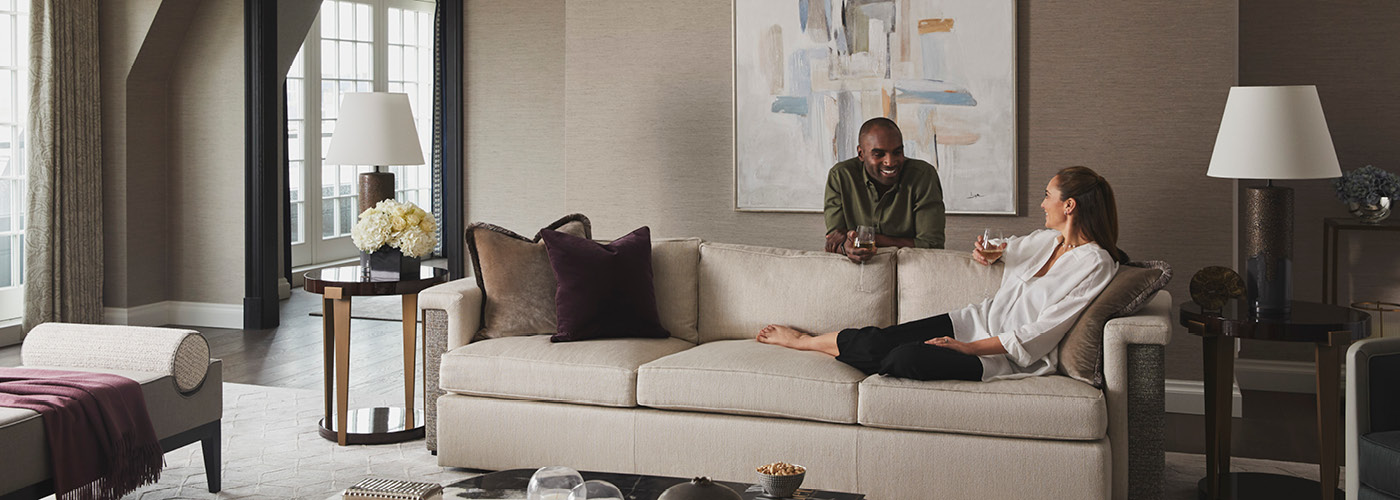 A woman lounges on a tan couch as a man leans on the back