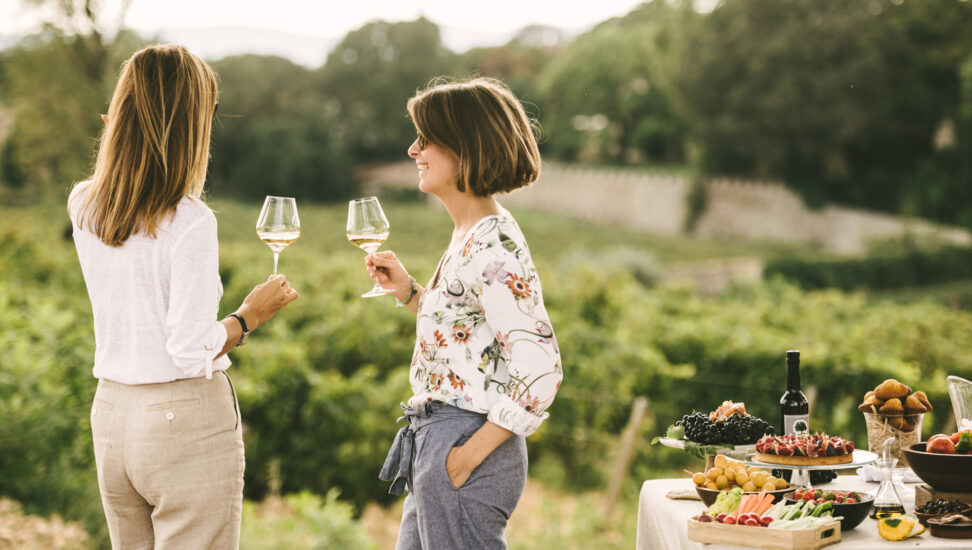 Two women drink a glass of white wine while standing among vineyards and private picnic
