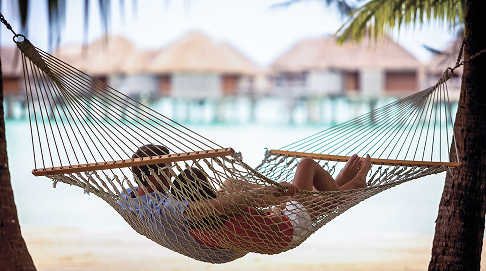 Couple lies in rope hammock between two palm trees, with beach, ocean and overwater bungalows in background