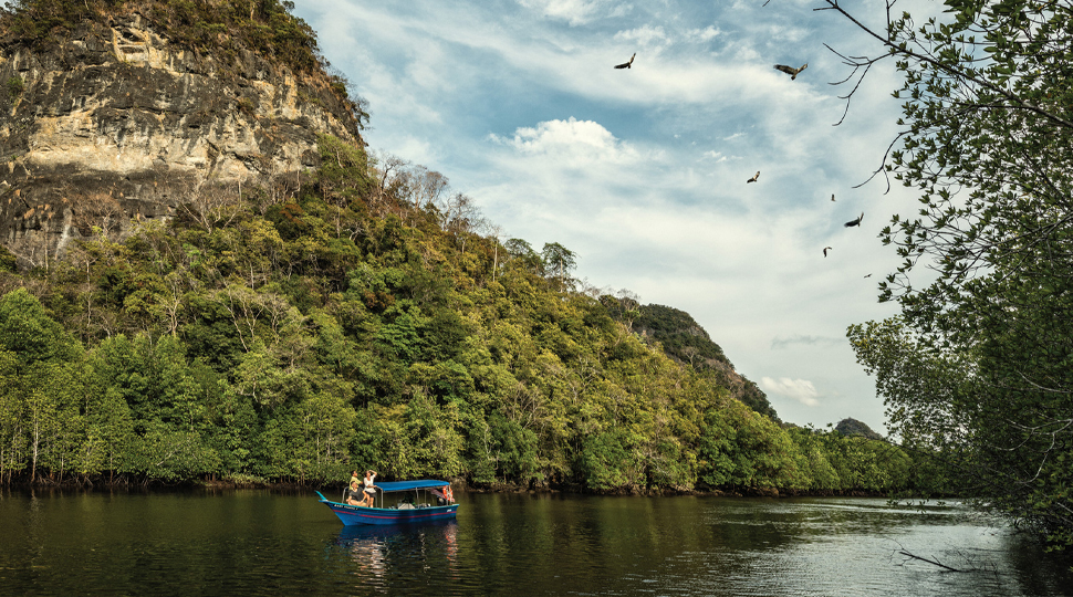 People on small blue boat on river under small rocky mountain, with eagles flying above