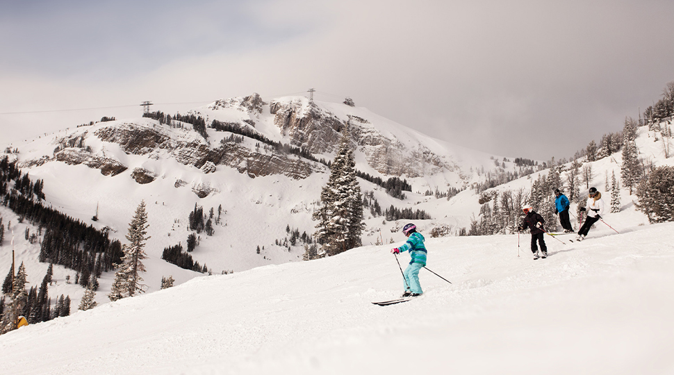 Four people wearing snow suits ski down a snow-covered mountain slope