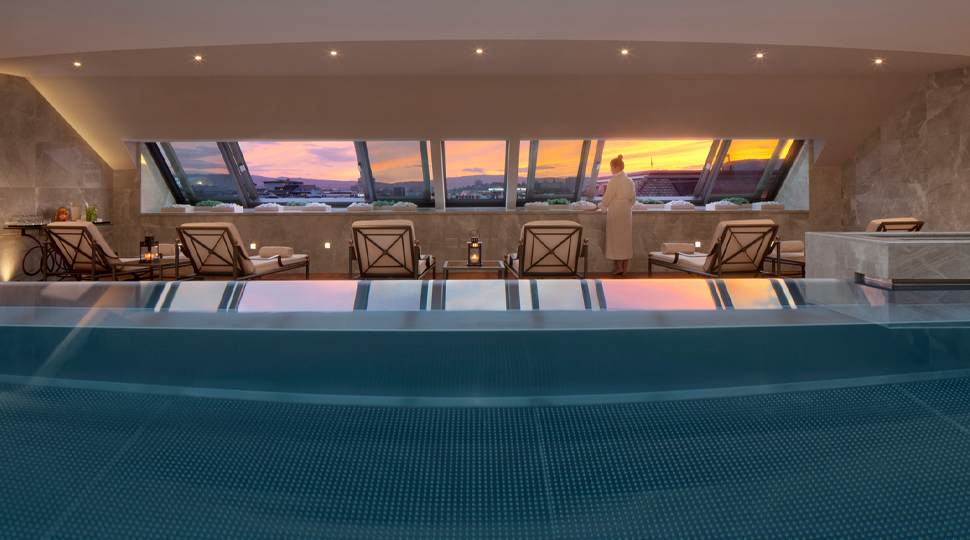 Indoor pool with lounge chairs on the side set beneath a slanted wall of windows