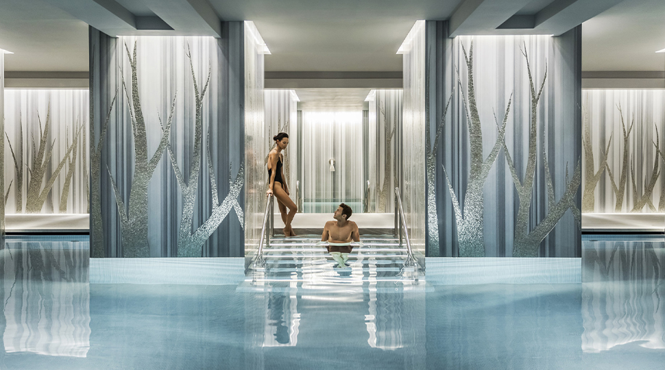 Man floats on steps of an indoor pool while woman stands next to him leaning on the hand rail