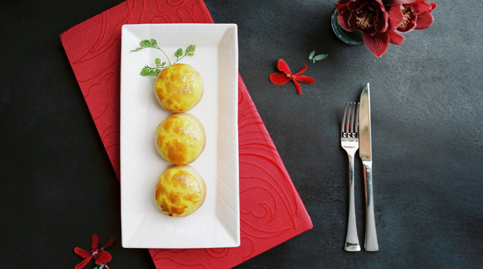 Three pork buns from Lung King Heen plated on a white rectangular dish with a spring of greenery and set on a black table with a bright red napkin and red flowers
