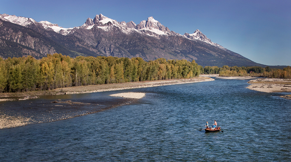 Person kayaking in a river with a tree line on the nearby shore and snow-capped mountains in the background