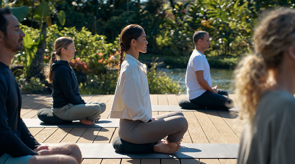 A group of five people sit on an outdoor wooden deck, each on their own yoga mat with their legs crossed in front of them