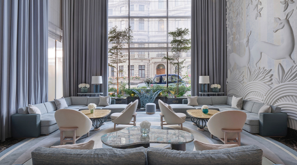 The lounge at Pavyllon London with light-blue furnishings, white arm chairs, light-wood coffee tables and sky-blue curtains framing double-height windows