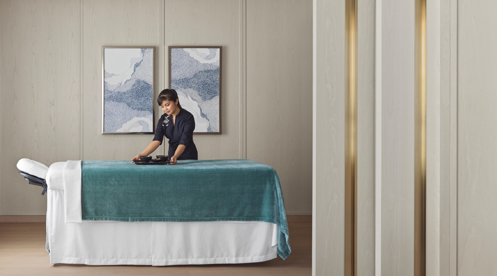 Spa therapist sets down a tray on a massage table draped with teal and white linens
