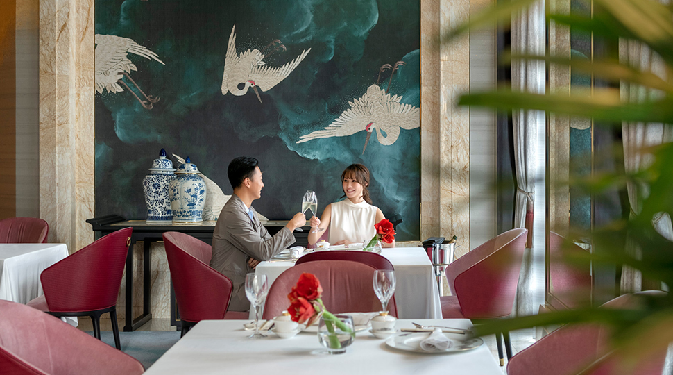 Two people sit at a table at Zi Yat Heen in Macao, featuring pink dining chairs, white-clothed tables and a mural wall displaying white birds on a dark teal background