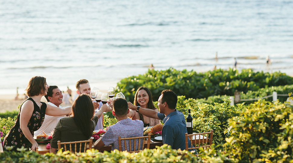 A group of seven people sit at a table set outdoors surrounded by greenery with the ocean in the background