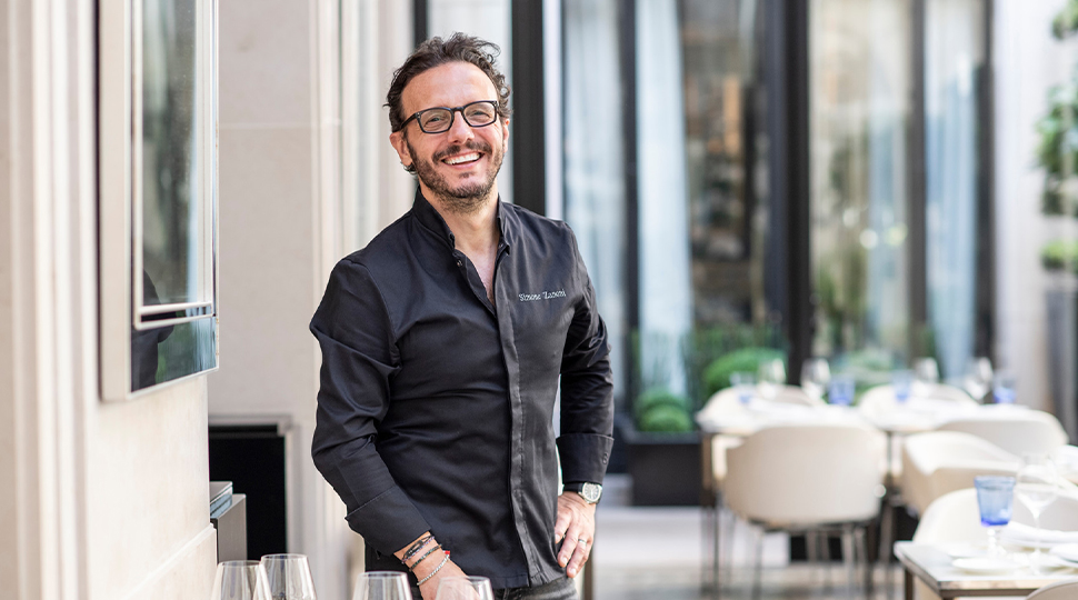 A smiling Chef Simone Zanoni stands on the Le George restaurant terrace wearing a black button-down shirt and glasses