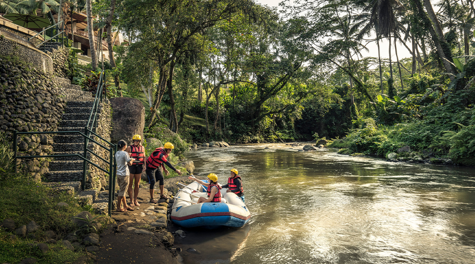Four people wearing orange life jackets board onto a white-water rafting boat docked on the side of a river in Bali