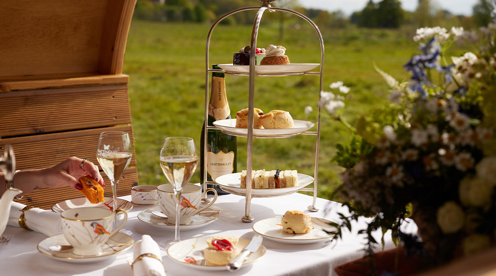 Three-tiered tray filled with pastries, various small plates, two wine glasses and a bottle of champagne are set on a white-clothed table in the middle of a meadow