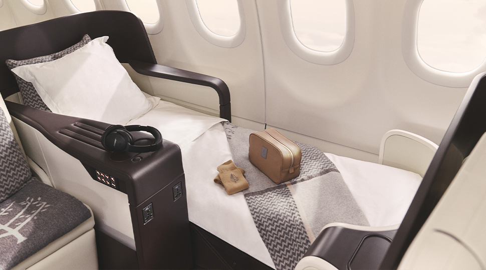 A seat on a plane is extended to a fully flat position with white linens, a grey throw blanket, slipper socks and a brown leather toiletry bag on top