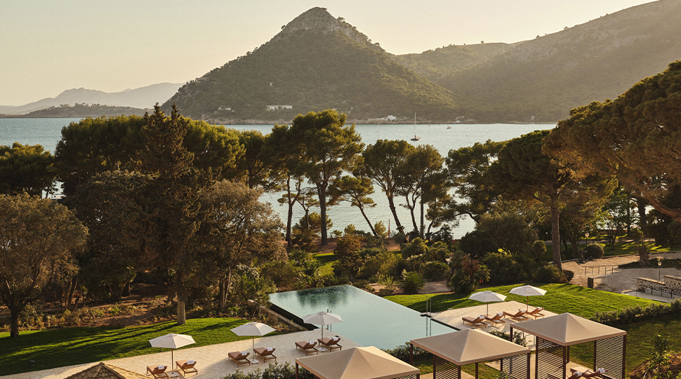 Rectangular outdoor pool with perpendicular pool deck sits in front of a treeline overlooking the ocean with mountains in the distance