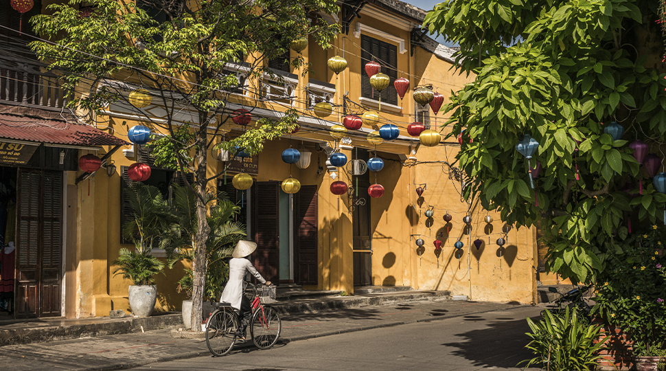 Person wearing silver robe, dark pants and Vietnamese conical hat rides a red bicycle in front of an orange building with hanging lanterns in front