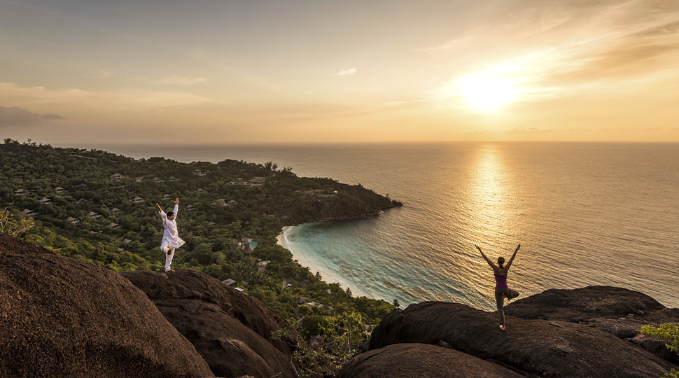 Two people on a cliffside overlooking the ocean, both in tree pose