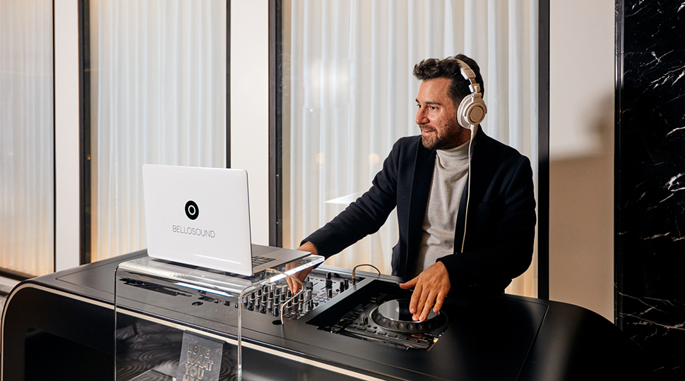Person wearing light-grey mock neck shirt and a black blazer sits behind a DJ stand wearing headphones