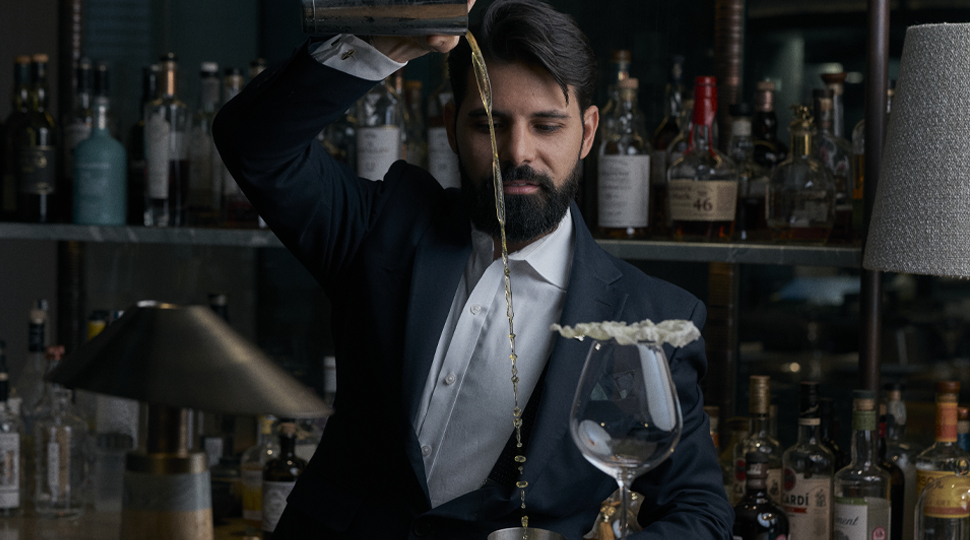 Bartender wearing a white collared shirt, no tie and a dark suit jacket pours a liquid from up high into a cocktail glass