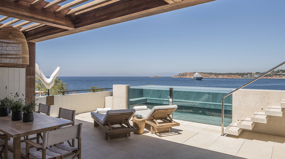 Open-air terrace with dining table, two lounge chairs and a glass pool overlooking the ocean