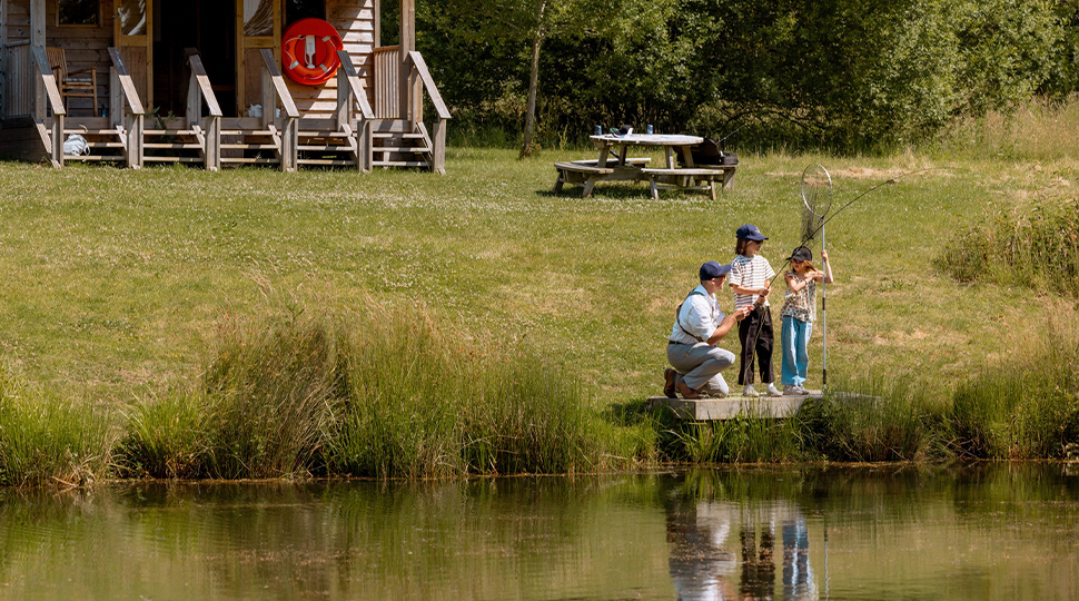 Two children and an instructor hold fly-fishing gear while standing by the edge of a pond