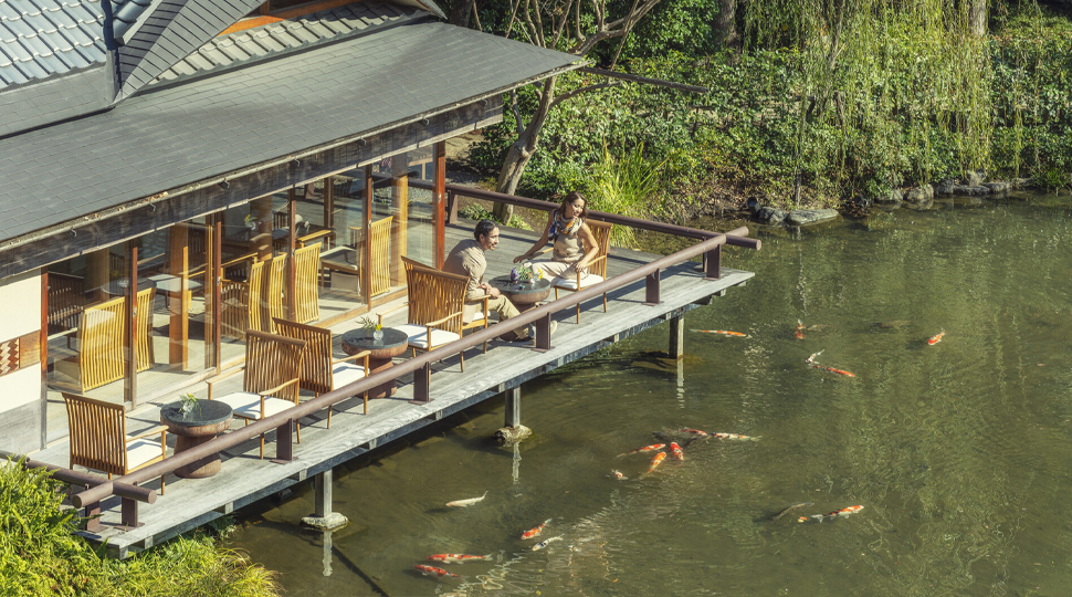 Two people sit on a restaurant terrace overlooking a pond with koi swimming in the water below