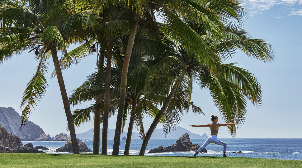 Person in a yoga pose on a lawn next to a collection of tall palm trees and the ocean in the background