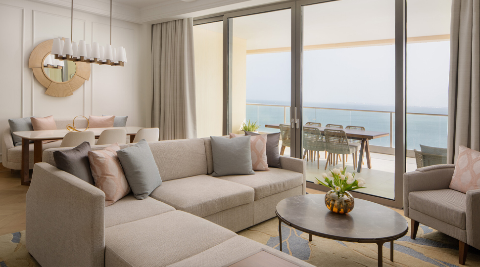 Living room with a light-beige, L-shaped sofa and round wooden coffee table in the centre, next to glass sliding doors that open up to a balcony overlooking the water