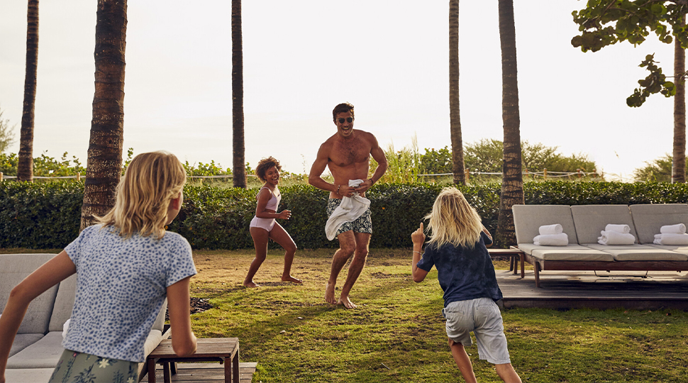 A parent runs around a palm-fringed yard with three young children