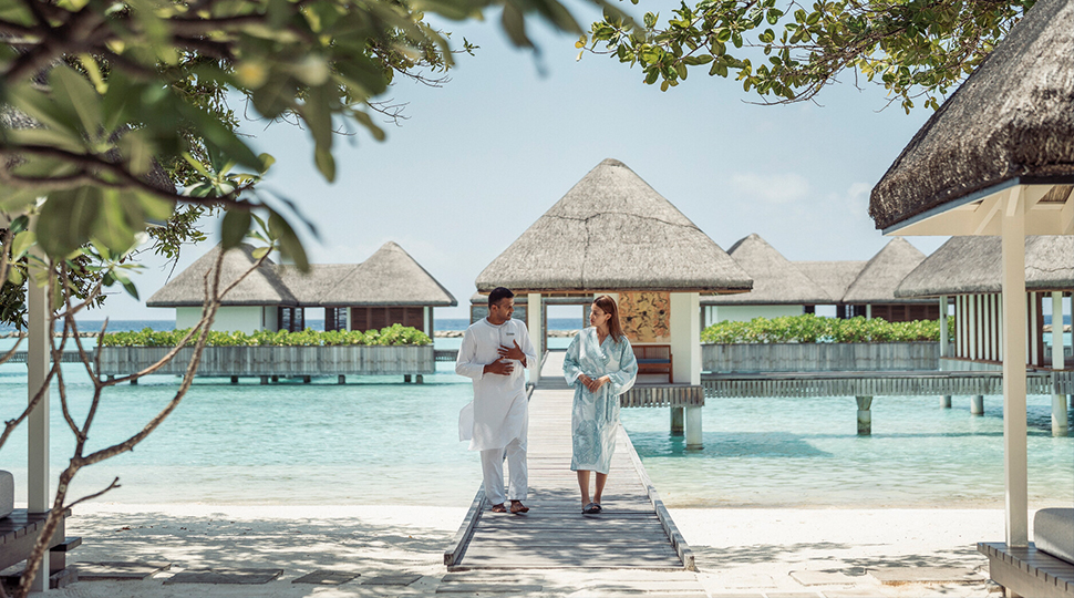 Two people walk along a narrow wooden dock that leads from the beach to an over-water pavilion