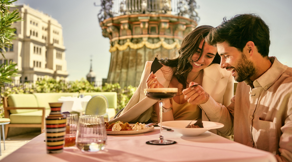 Two people smile as they share a dessert at a table at a rooftop restaurant