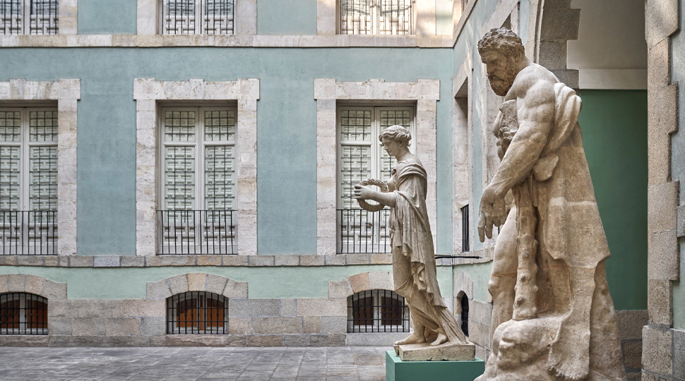 Two marble statues stand in a plaza surrounded by a light-blue building