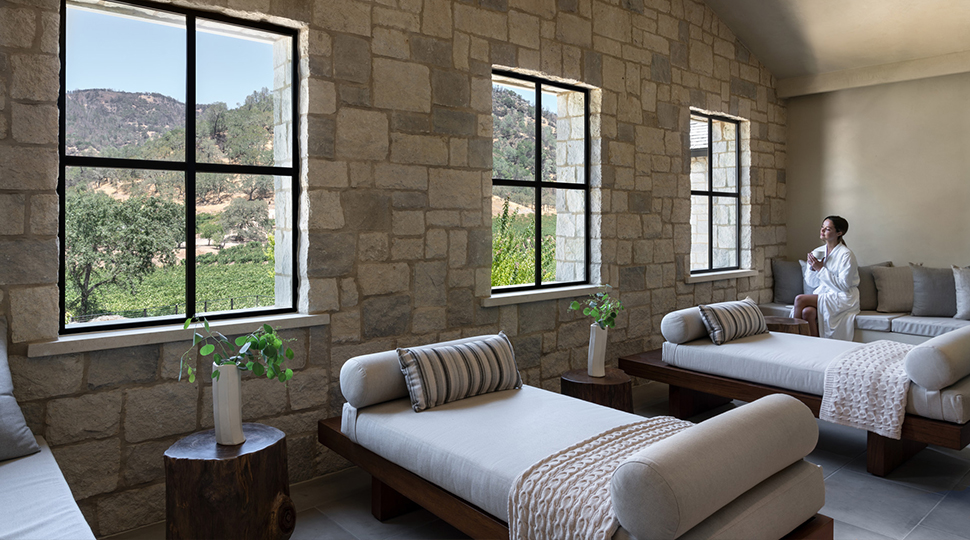 A person wearing a white spa robe sits sipping a cup of tea while looking out the window next to a row of daybeds set along a stone wall