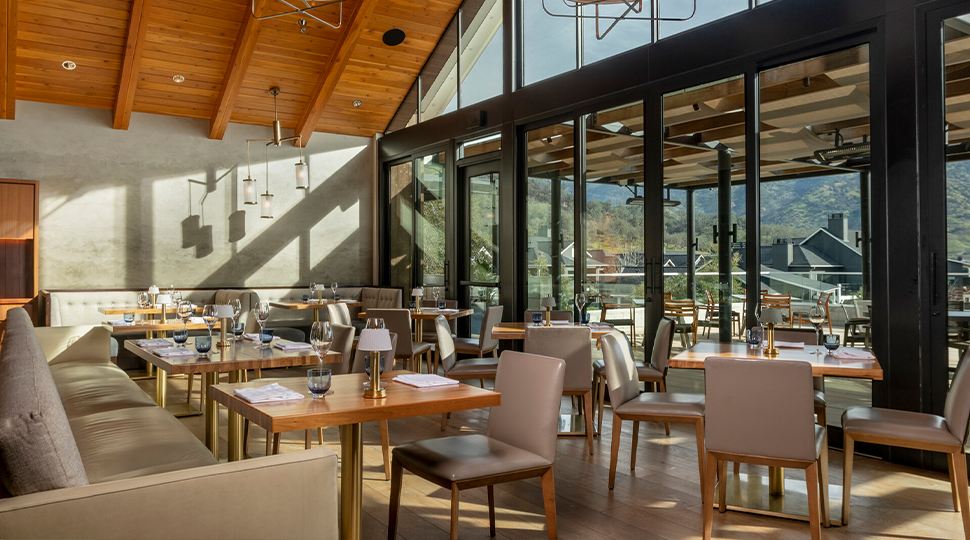 Interior restaurant dining room filled with square wooden tables for four, a pitched wooden ceiling, and a wall of windows and glass doors leading out to a covered terrace